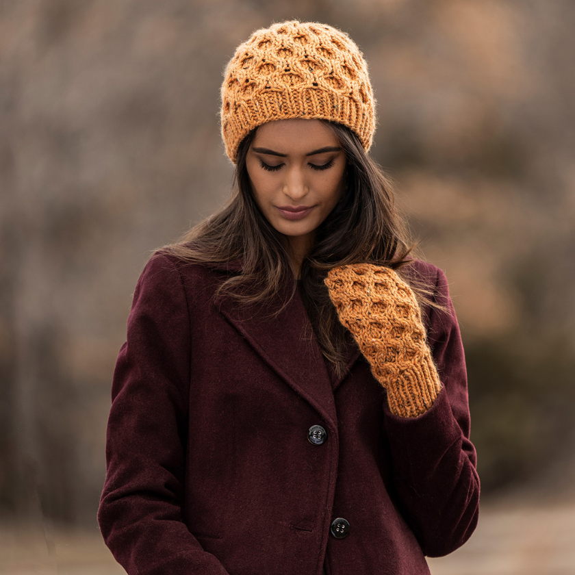 Woman wearing a maroon coat, mustard Huntington knit hat, and matching mittens against a blurred natural background.