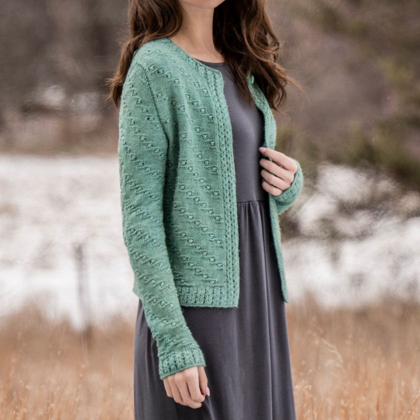 A model shows off the arm detail of the Ocean Bloom Cardigan over a gray dress, standing outdoors with a trees in the background.