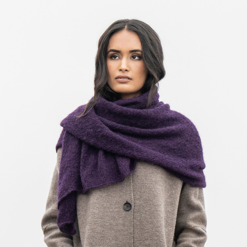 A woman wearing a gray coat and an Elderberry Stardust Wrap scarf-style around her neck, standing in a natural outdoor setting with trees in the background.