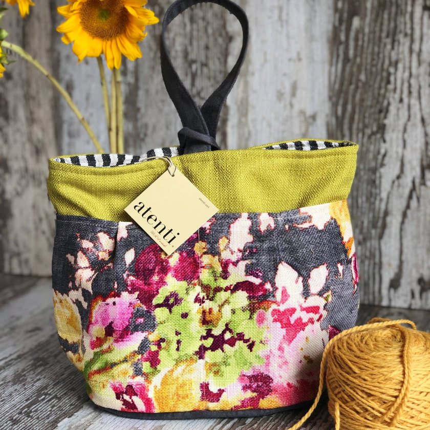 Floral-patterned fabric knitting basket with an atenti tag on a wooden surface, next to a ball of yarn and sunflower.