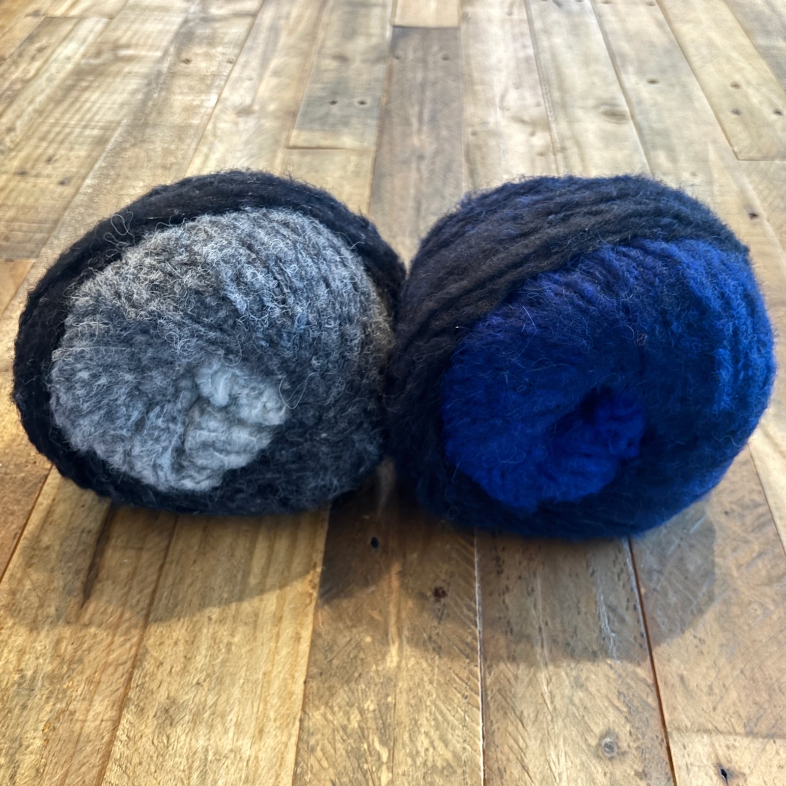 Two skeins of super bulky Piuma Alpaca Degradé yarn in Greys and Blues for the Degradé Drift Scarf on a wooden background