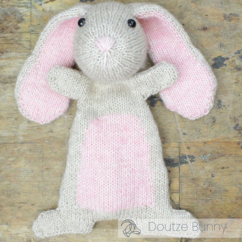 Knitted bunny toy with pink ears and body on a wooden surface, branded 'Doutze Bunny'.