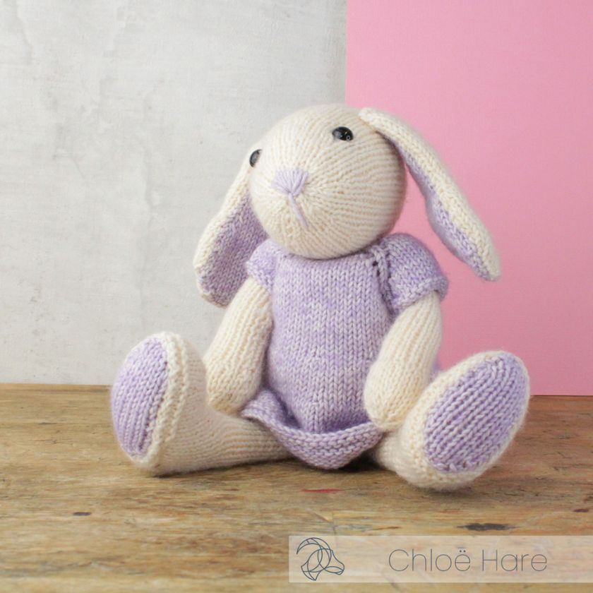Knitted Amigurumi toy bunny in a purple dress on a light gray and pink background