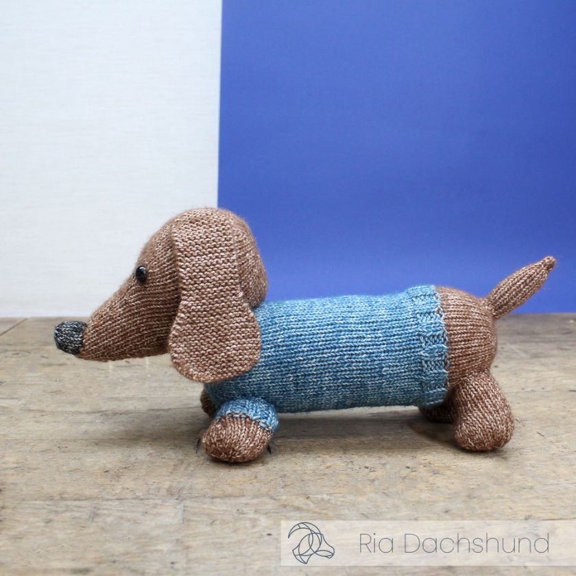 Knitted dachshund toy from Amigurumi wearing a blue sweater on a wooden surface with a blue and white background.