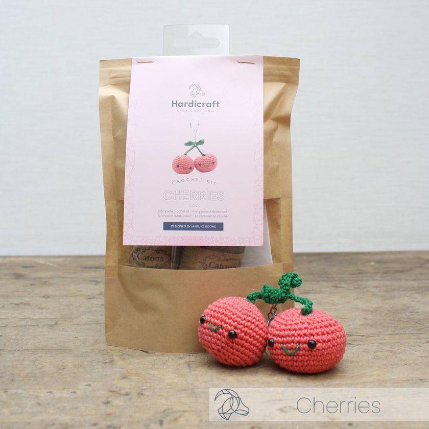 Two pink crochet cherries with green stems in front of the crochet kit packaging from 'Hardicraft'.