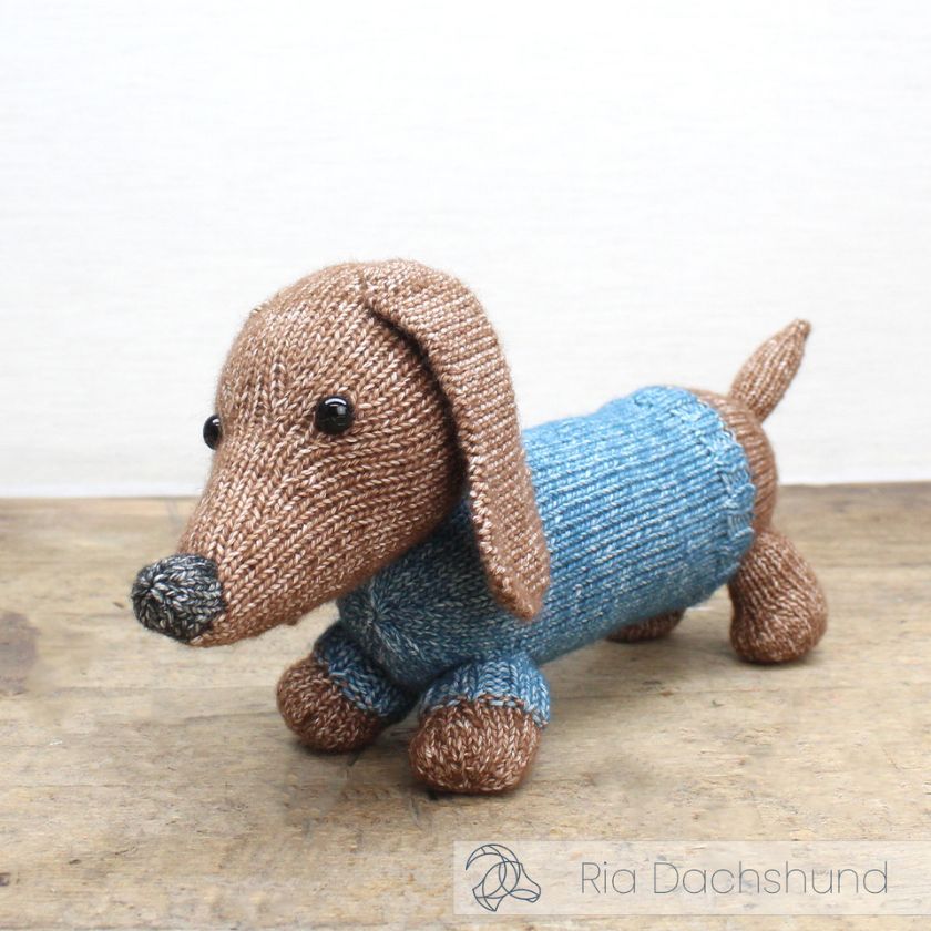Knitted dachshund toy from Amigurumi wearing a blue sweater on a wooden surface with a blue and white background.