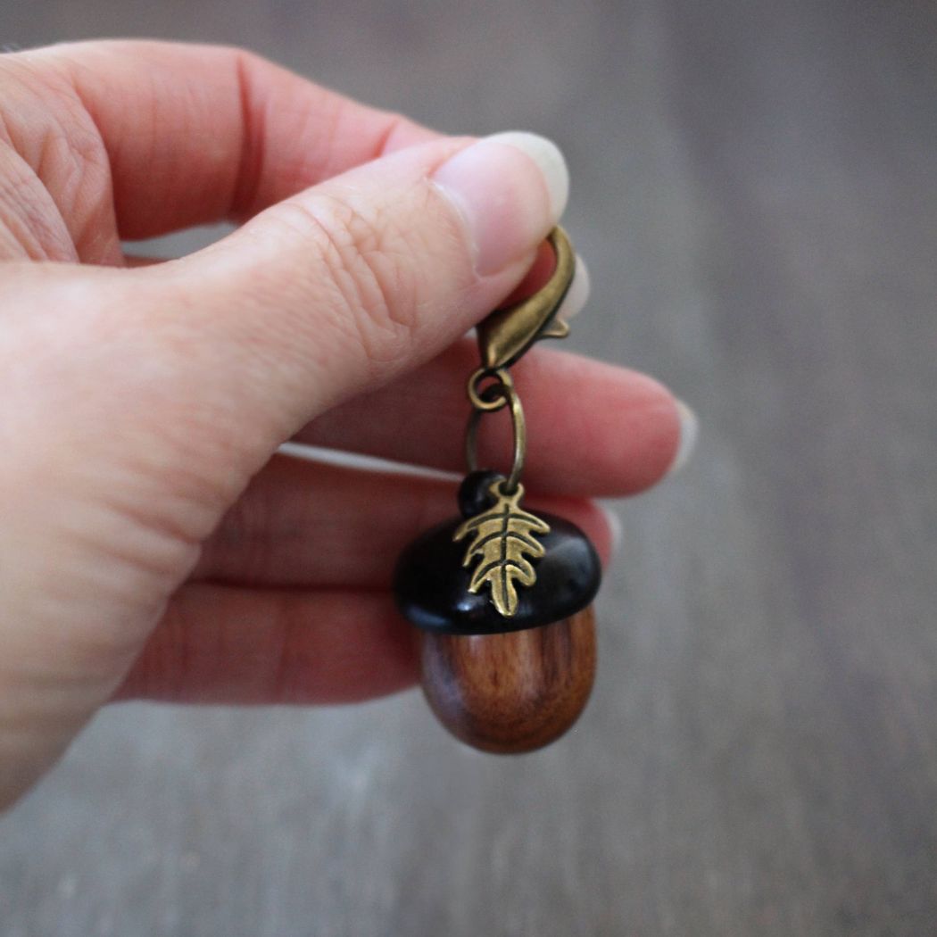 Hand holding a wooden acorn stitch marker holder with a leaf design on a neutral background