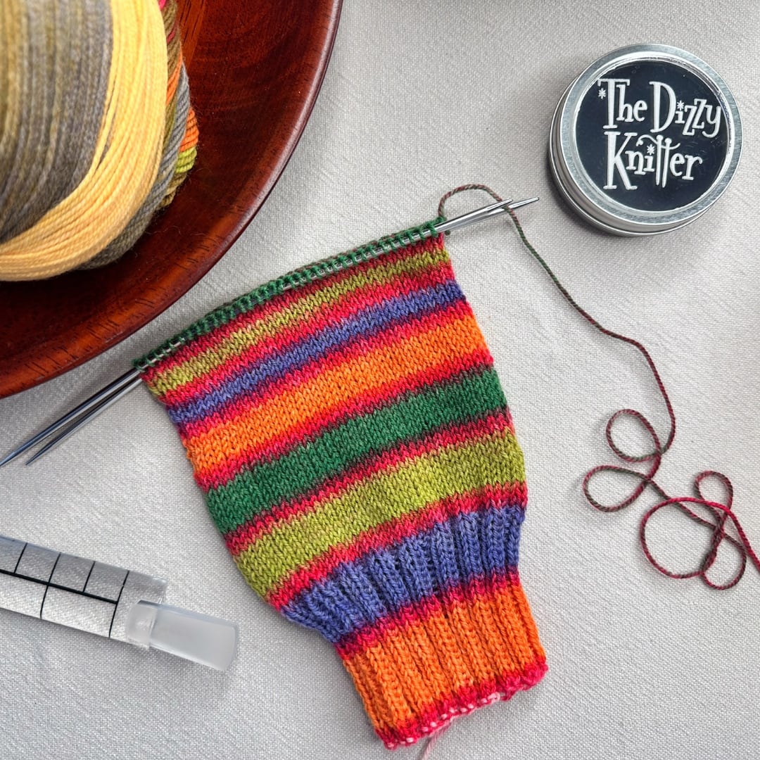 Colorful striped knitted Laine Dunord socks on needles with yarn and knitting tools on a light surface.