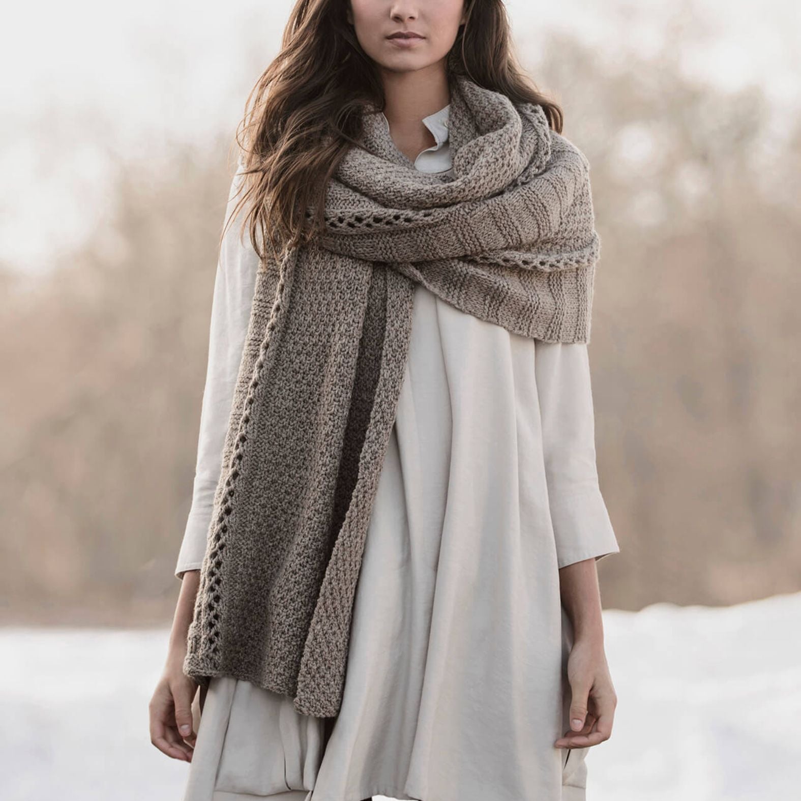 Model wearing the Endless Wrap knit scarf in a neutral wool color, styled loosely around the shoulders with soft textured stitch details visible.