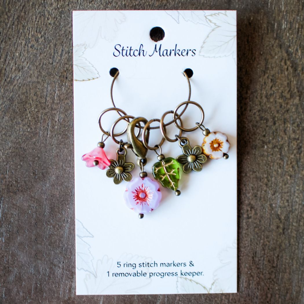 Set of spring themed stitch markers on a card with floral designs against a wooden background
