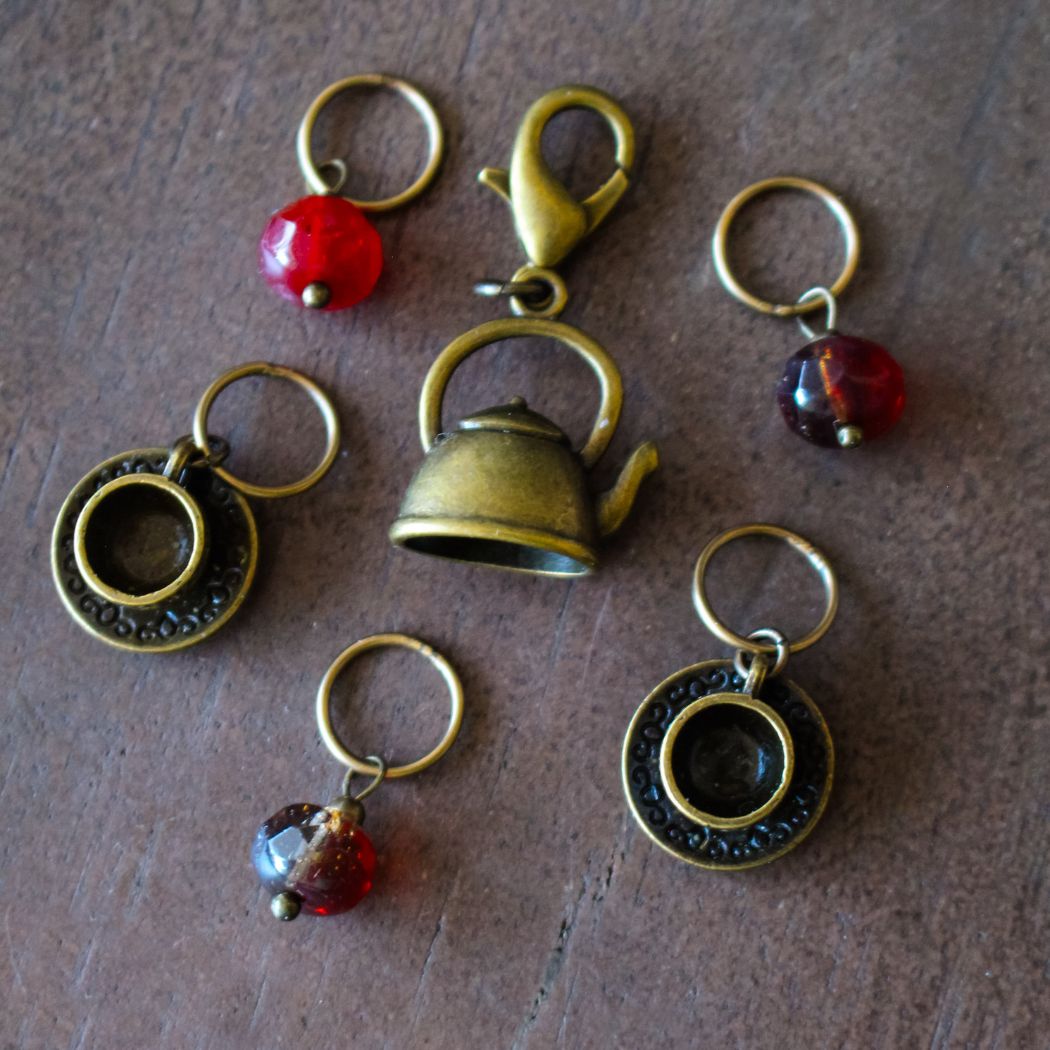 Decorative stitch markers with tea themed charms on a wooden surface.