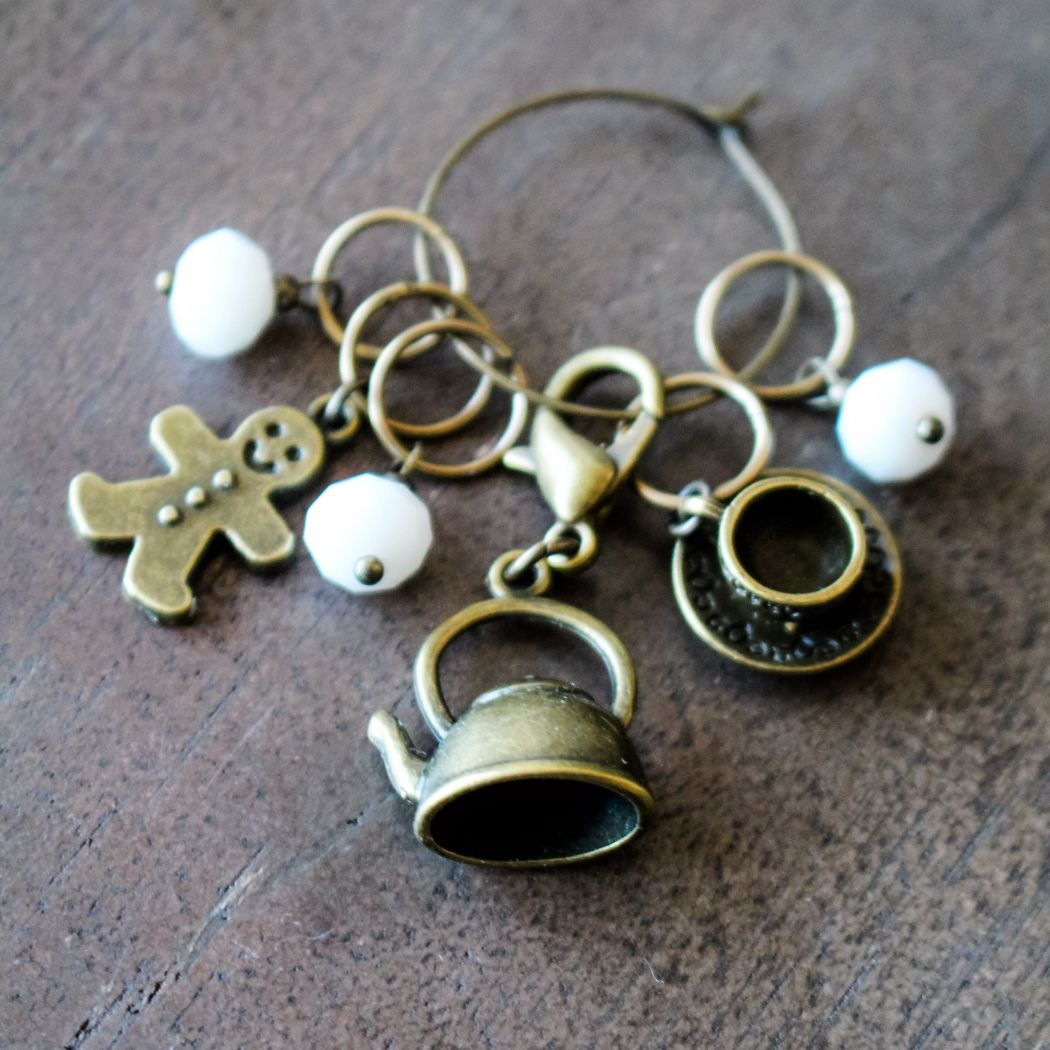 Winter themed stitch markers with a teapot, gingerbread man, and white beads on a wooden surface.