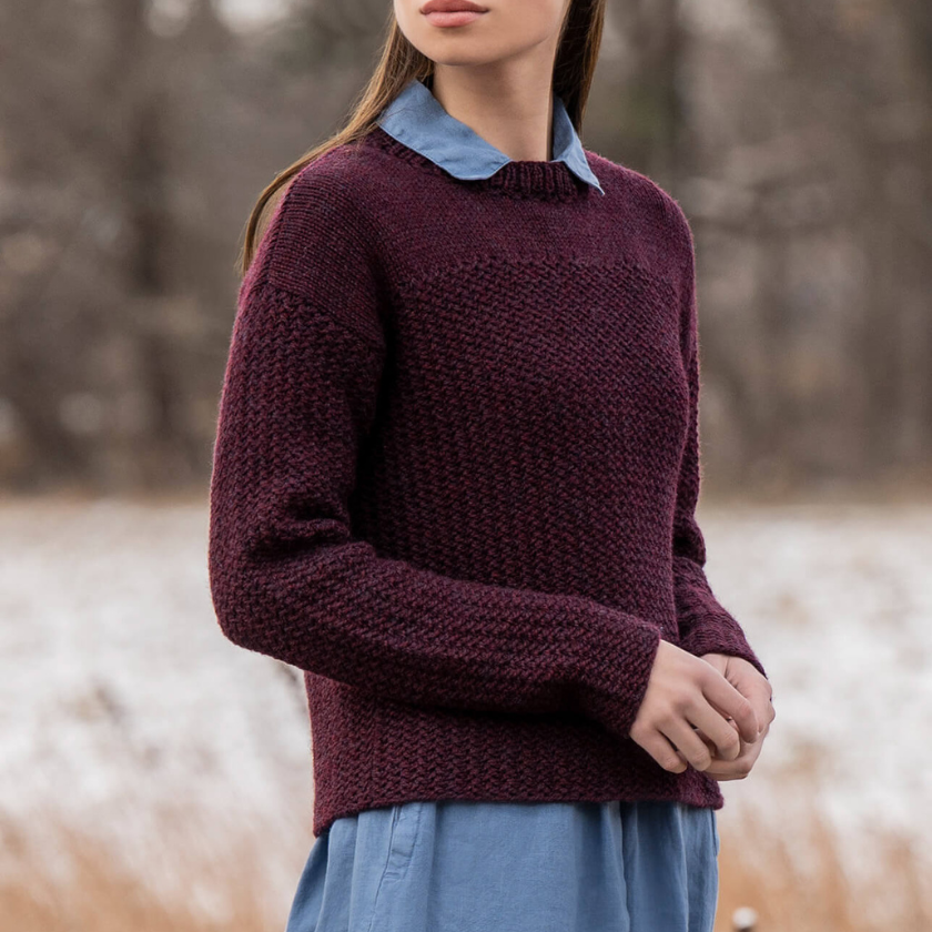 Person wearing a maroon knitted Park Avenue pullover over a blue shirt in a natural setting