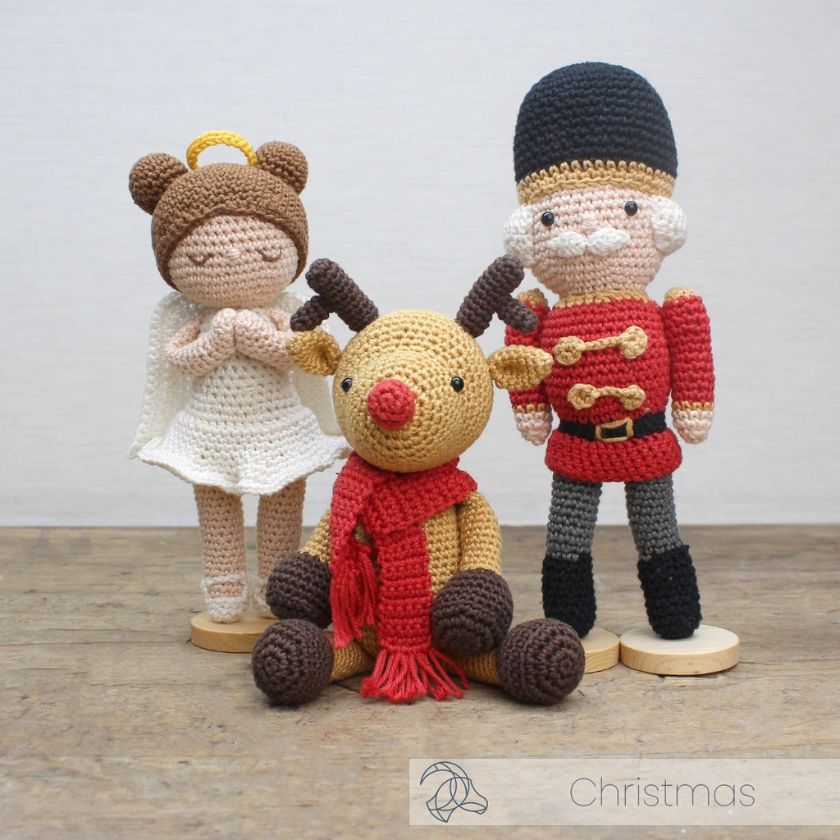 Three crochet Christmas amigurumi characters including a reindeer, angel, and soldier on a wooden surface.