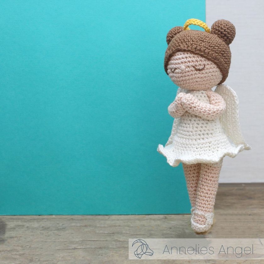 Crocheted angel doll with a brown hair, halo, and white dress against a teal and white background.