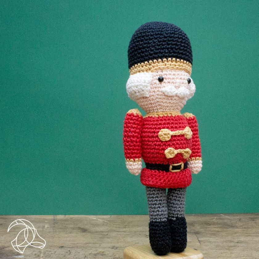 Crocheted toy figure of a guard in red uniform with a black hat on a wooden base against a teal background.