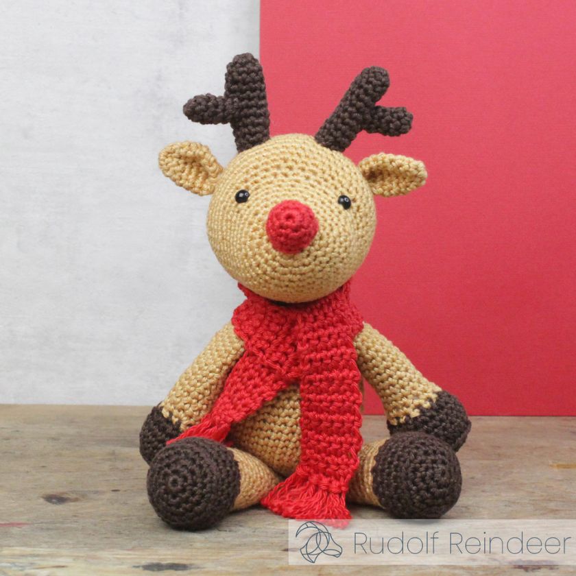 Crocheted reindeer toy with a red scarf on a wooden surface against a white and red background.